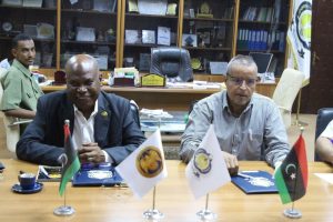BFCE666C-C2C5-4CC1-BEDA-8CB46F6DC40D - جامعة سبها | Sebha University Two men sit at a conference table with flags and documents in an office setting.