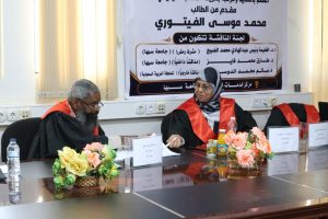 Two panelists in black robes with red sashes sit at a conference table in a meeting room, with a large Arabic banner and pink floral centerpieces behind them.