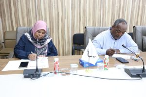 Two professionals sign documents at a conference table, with microphones and a small flag between them.