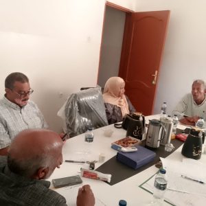 CD7A2FAE-2C61-4FC7-AE9E-B5CB90F870D5 - جامعة سبها | Sebha University Four adults sit around a conference table with papers, pens, water bottles, and snacks in a casual meeting room. A woman wearing a hijab is present.