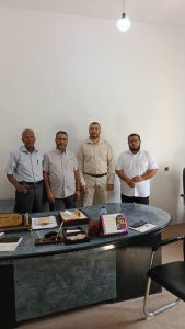 D0490DBA-5522-4B09-863A-D82A2E1FF59D - جامعة سبها | Sebha University Four men in light shirts pose for a group photo in a neutral office behind a desk.