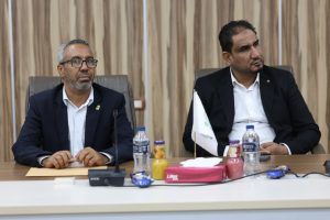 D6A5B65C-4AC7-4012-BA2F-D320115D0113 - جامعة سبها | Sebha University Two men in suits sit at a conference table with microphones and papers, looking attentive in a formal meeting room.