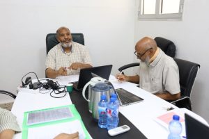 Two men sit at a white conference table with laptops, papers, and water bottles, discussing work.