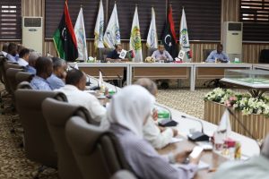 Formal meeting in a conference room with a row of flags behind the head table and attendees seated around curved desks.