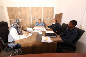 D9BDC1A6-C264-4A8B-BAA3-561829B52D98 - جامعة سبها | Sebha University Four colleagues sit around a wooden desk in a small office, reviewing papers and documents collected on the table. A woman wearing a hijab is on the left; three men sit across and to the right, one wearing a face mask. Behind them are wooden cabinet panels and a stack of boards.