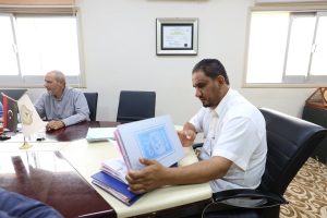 Two men sit at a polished conference table in an office, one flipping through a stack of documents as they work at a meeting.