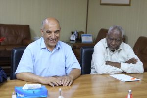 Two men sit at a conference table, one smiling to the camera while the other looks serious during a meeting.
