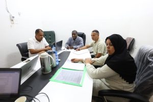 E52FBAB9-858B-4F12-97C7-153D68237EEE - جامعة سبها | Sebha University Five colleagues sit around a conference table in a bright room, reviewing documents with laptops and beverages on the table.