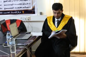 Male graduate in a black gown with a yellow hood sits at a desk, reading a notebook beside two laptops and water bottles on the table.