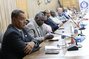 EB8D1E8F-2412-48B2-9CB1-8CD3C7A0F518 - جامعة سبها | Sebha University A line of men in suits seated at a long conference table with microphones, notes, and water bottles during a formal meeting.