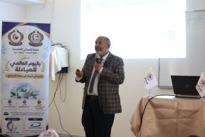 EC02EDDE-4474-432D-9CED-A1E1451DB1AC - جامعة سبها | Sebha University Presenter in a plaid blazer speaks into a microphone at a seminar, with a large banner on the left and a laptop on a table to the right behind him.