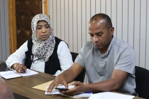 ECA26147-6648-473D-A7C1-969CAD2642A1 - جامعة سبها | Sebha University Two adults seated at a table in a meeting; the woman in a patterned hijab is writing in a notebook while the man in a gray shirt looks at his phone and papers are on the table.
