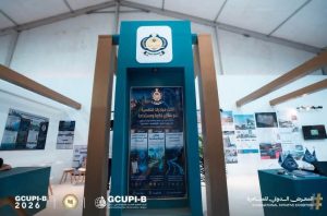 Teal exhibition booth with a tall central pillar displaying a logo and a large Arabic poster inside a white tent.