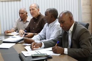 Four men sit at a conference table signing and reviewing documents during a business meeting in a boardroom setting.] ,"