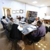 Group of professionals seated around a conference table in a meeting room, actively discussing documents and notes:**