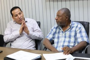 F8F3EE18-6591-4335-B83A-7E4621951407 - جامعة سبها | Sebha University Two men in a meeting at a conference table; one speaks with hands clasped, the other listens with a pencil in hand.