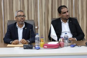 FA4531D0-B313-47F1-8F53-61F5C929A3DC - جامعة سبها | Sebha University Two men in suits sit at a conference table with microphones, water bottles, and a small flag in front of them, during a formal meeting