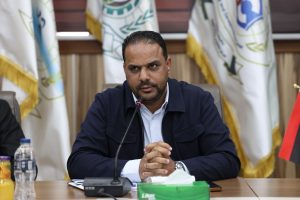 FED54325-7E90-4E5B-A094-FD4D8B9B7E4A - جامعة سبها | Sebha University Man in a white shirt and dark jacket sits at a conference table, hands clasped, speaking into a microphone with flags in the background.