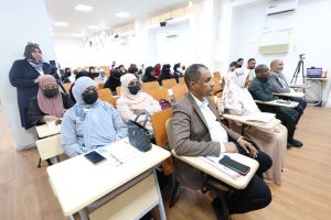 Audience in a bright lecture hall; women in hijabs sit in front rows, a man in a brown blazer at a desk with notebooks and phones visible.