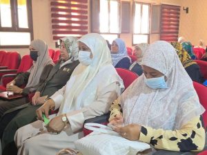Group of women in hijabs and masks sitting in red auditorium chairs at a conference.