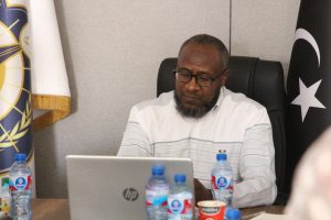 Man with glasses and a beard sits at a desk using an HP laptop, with flags in the background of a office setting.