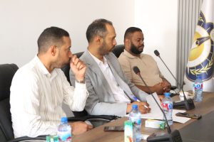 Three men seated at a conference table with microphones and papers, engaged in a panel discussion, with bottled drinks on the table and a flag in the background.
