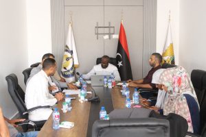 Group of people seated around a conference table in a meeting room, with flags in the background behind the speakers.