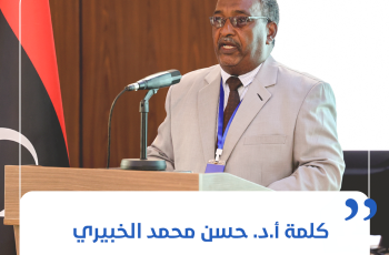 Man in a light gray suit giving a speech at a wooden podium with a microphone and a blue lanyard, in front of a flag backdrop.
