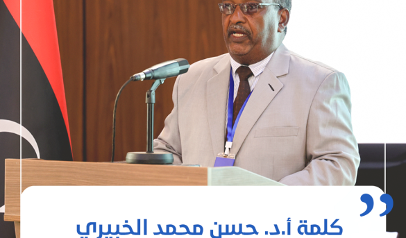 inbound1873104467690712026 - جامعة سبها | Sebha University Man in a light gray suit giving a speech at a wooden podium with a microphone and a blue lanyard, in front of a flag backdrop.