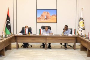 inbound2357309040045849704 - جامعة سبها | Sebha University Three men sit at a U-shaped wooden conference table in a formal meeting room, with flags and papers, a desert arch image on the screen behind them.