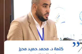 Man in a beige suit with a blue lanyard speaking at a conference, microphone in front of him