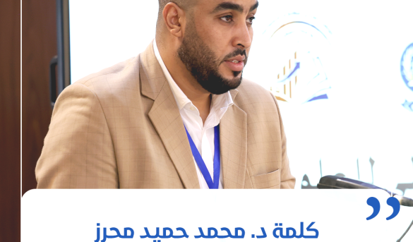 inbound2698607400408530554 - جامعة سبها | Sebha University Man in a beige suit with a blue lanyard speaking at a conference, microphone in front of him