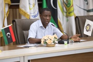 inbound2701926445557602972 - جامعة سبها | Sebha University Man in a light shirt speaks into a microphone at a conference table, with flags and a flower centerpiece behind him.