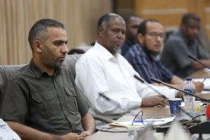 A man in a dark green shirt sits at a conference table with others in the background, listening attentively during a meeting.