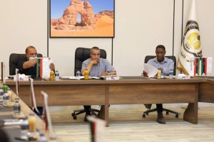 inbound2794590683024011766 - جامعة سبها | Sebha University Three men sit at a long conference table in an office, reviewing papers and beverages on the desk, with Jordan flags and a large flag in the corner. A desert landscape photo hangs on the wall behind them.