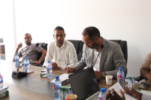 Group of men seated around a conference table in a meeting room, with microphones, notebooks, and water bottles on the table.
