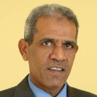 Portrait of a middle-aged man in a dark suit and blue shirt against a yellow background.