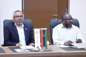 inbound3254387982476624388 - جامعة سبها | Sebha University Two men sit at a conference table with a flag display, notebooks and a laptop nearby.