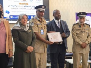 Officials in uniforms and a suit present a certificate at an award ceremony, with a banner in the background.