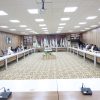 A large conference room with participants seated along U-shaped tables and flags displayed at the back wall.