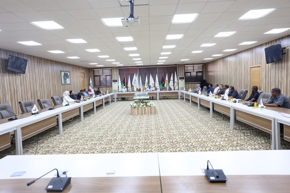 A large conference room with participants seated along U-shaped tables and flags displayed at the back wall.