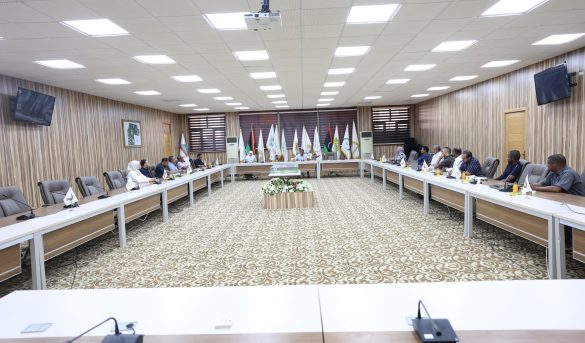 inbound3741884239959556750 - جامعة سبها | Sebha University A large conference room with participants seated along U-shaped tables and flags displayed at the back wall.