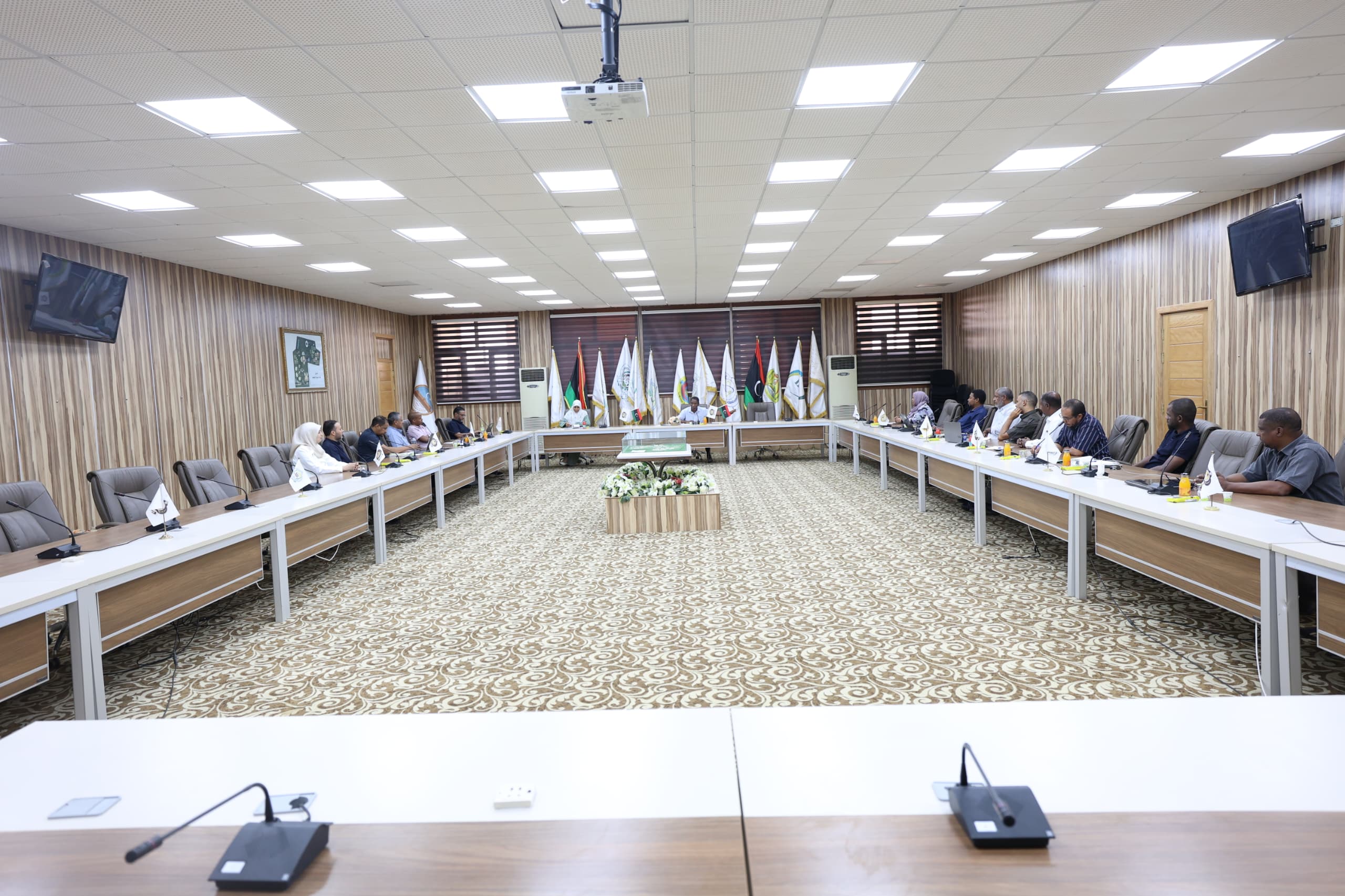 A large conference room with participants seated along U-shaped tables and flags displayed at the back wall.