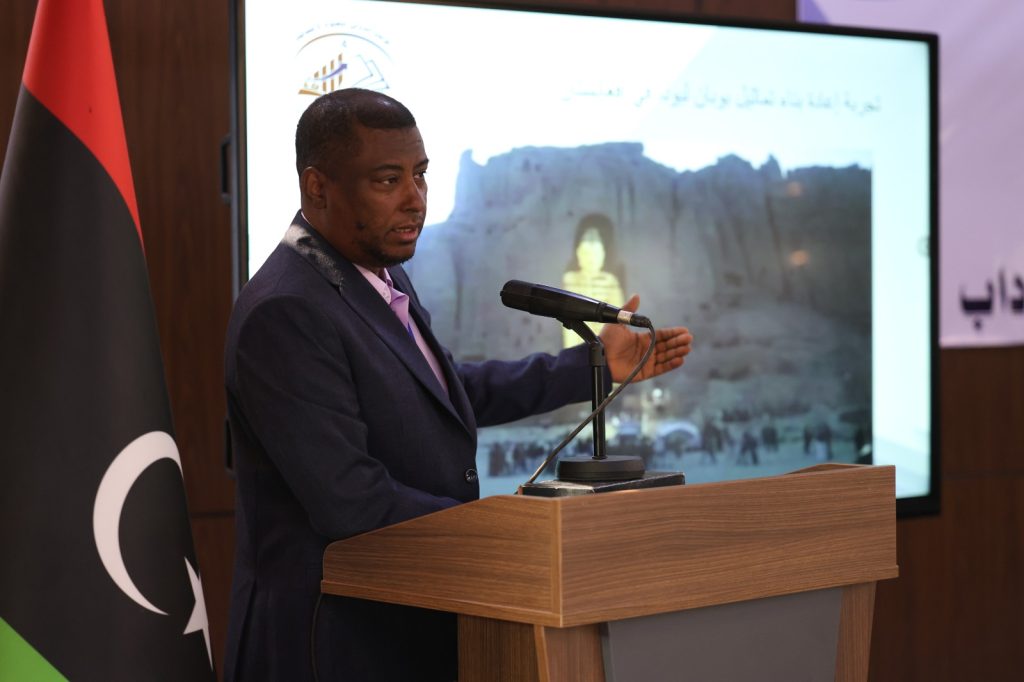 Man in a dark suit speaks at a wooden podium with a microphone, facing an audience; a flag stands nearby and a large screen shows a backdrop image with Arabic text.