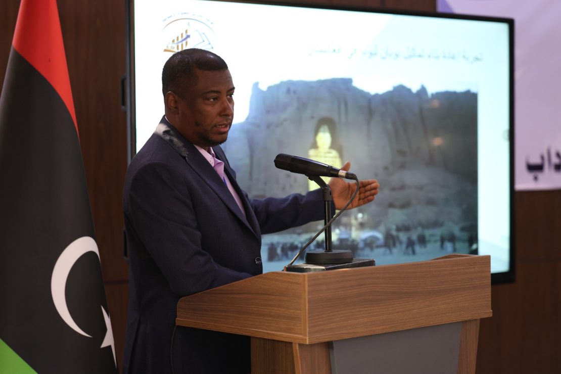 Man in a dark suit speaks at a wooden podium with a microphone, facing an audience; a flag stands nearby and a large screen shows a backdrop image with Arabic text.
