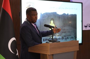 Man in a dark suit speaks at a wooden podium with a microphone, facing an audience; a flag stands nearby and a large screen shows a backdrop image with Arabic text.