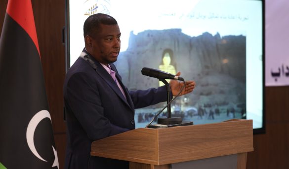 inbound383230624907863628 - جامعة سبها | Sebha University Man in a dark suit speaks at a wooden podium with a microphone, facing an audience; a flag stands nearby and a large screen shows a backdrop image with Arabic text.