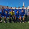 Group of young soccer players in blue jerseys pose for a team photo on a green field with buildings and a clear blue sky in the background, front row kneeling.