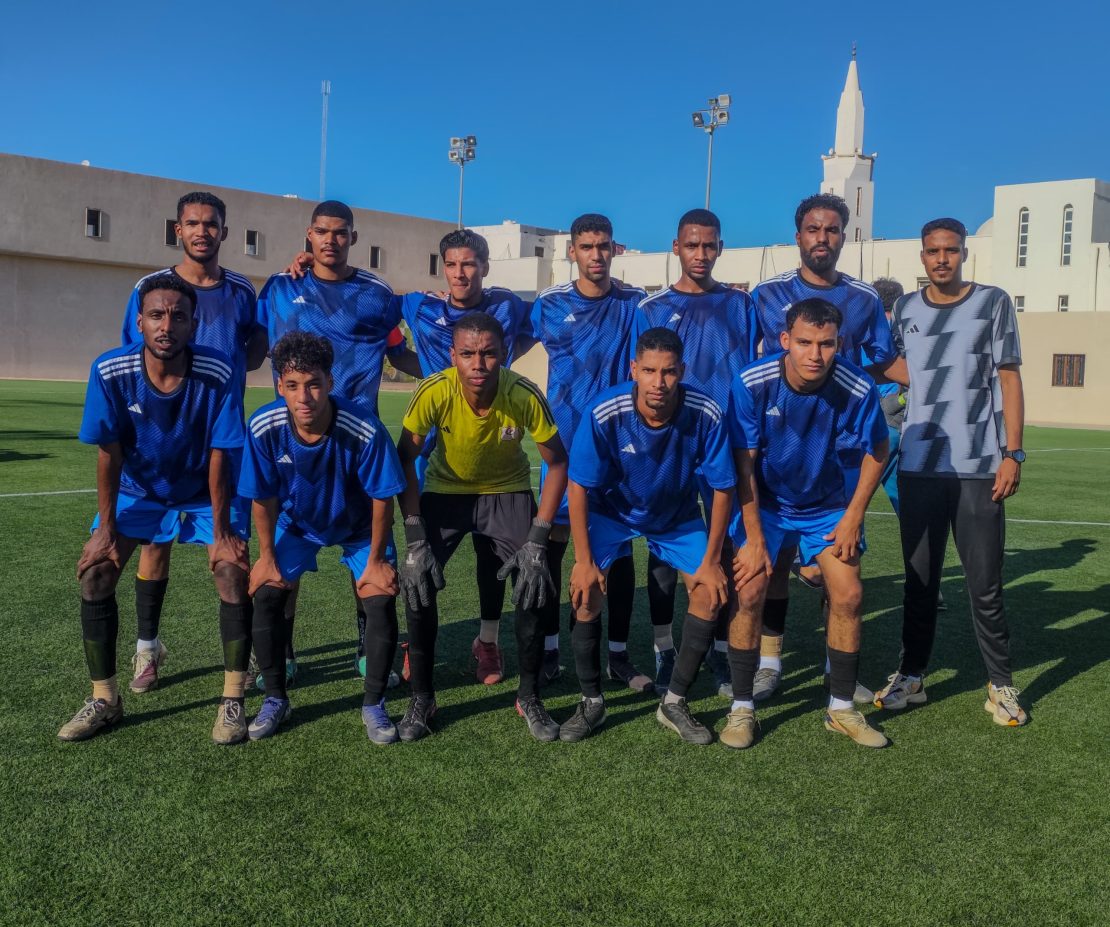 Group of young soccer players in blue jerseys pose for a team photo on a green field with buildings and a clear blue sky in the background, front row kneeling.