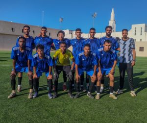 inbound3955271721915703340 - جامعة سبها | Sebha University Group of young soccer players in blue jerseys pose for a team photo on a green field with buildings and a clear blue sky in the background, front row kneeling.